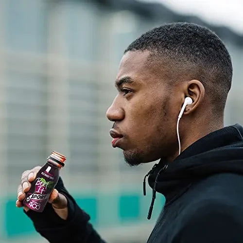 Man holding a bottle of BEET IT beetroot juice with earphones in his ears, standing outdoors.